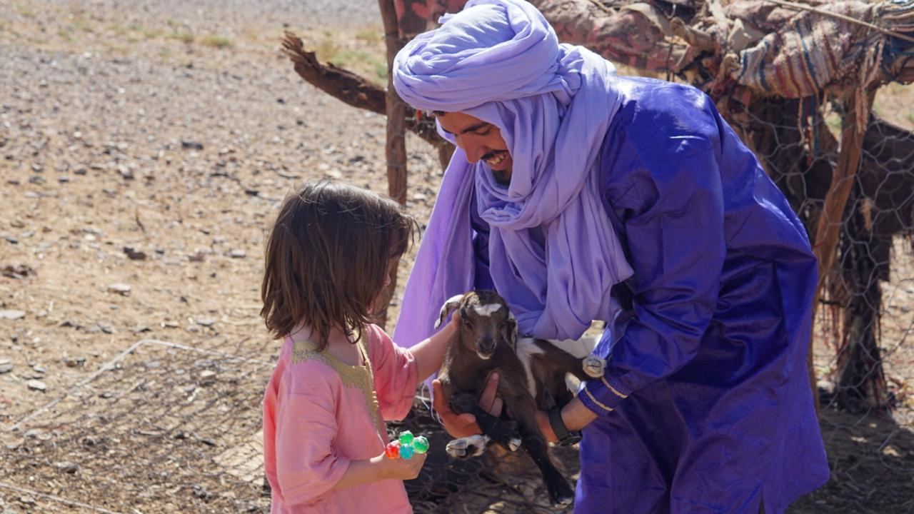 A Berber guide introducing a young traveler to a baby goat near a desert camp