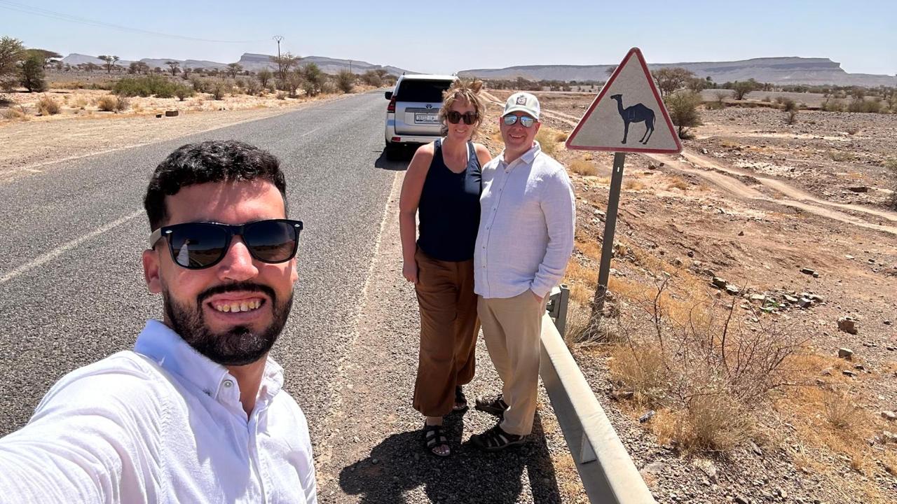 Mubarak with a group of travelers on the road, next to a camel-crossing sign